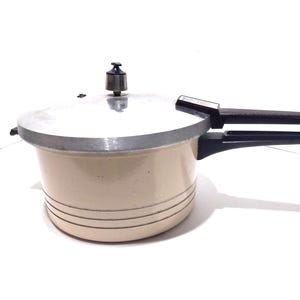 Könnte beinhalten: Cremefarbener Schnellkochtopf mit silbernem Deckel und schwarzen Griffen. Der Topf hat drei horizontale Linien, die um den Boden geätzt sind. Ein silbernes Ventil sitzt auf dem Deckel. Der Griff erstreckt sich horizontal von der Seite.