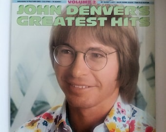 May include: A vintage vinyl record cover featuring a smiling John Denver with brown hair and glasses. The cover is white with green text that reads "John Denver's Greatest Hits, Volume 2".