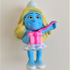 Puede incluir: Una figurita de Pitufo azul con cabello rubio, usando un sombrero blanco y un vestido blanco, sosteniendo una caja de regalo rosa con una cinta rosa.
