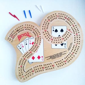 May include: Wooden cribbage board in a heart shape with the Bicycle logo and playing cards. Includes red, blue, and white pegs. The board has a spiral track with numbered holes for scoring.
