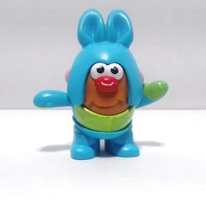May include: A colorful toy figure resembling a potato with a blue bunny-ear hat, arms, and legs. It has a tan face, a red mouth, and large white eyes. The figure also has a green belly band and a green hand.