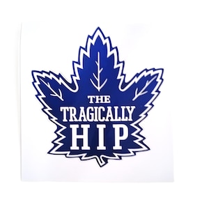 Puede incluir: Adhesivo azul y blanco con un diseño de hoja de arce estilizada. El texto "THE TRAGICALLY HIP" está centrado. El adhesivo tiene un fondo blanco, un homenaje a la banda canadiense.