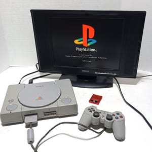 May include: A gray Sony PlayStation game console with a top-loading disc drive, a matching gray wired controller, and a small red memory card. The console is connected to a black Daenyx monitor displaying the PlayStation logo and startup text.