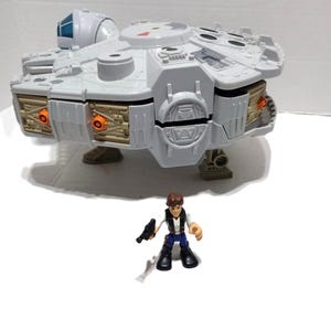 May include: A gray plastic toy spaceship with a circular body, a side cockpit, and tan side panels with orange circular accents. The spaceship stands on three legs. In front of the ship stands a small plastic figure with brown hair, a white vest, blue pants, and black boots, holding a small black toy blaster.