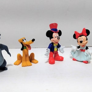 Puede incluir: Colección de cinco figuras de personajes de Disney. Incluye a Baloo el oso, Pluto el perro, Mickey Mouse con sombrero de copa, Minnie Mouse con vestido y Simba el cachorro de león. Cada figura es de colores brillantes y se mantiene de pie.