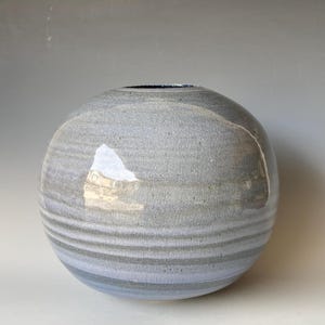 May include: A spherical ceramic vase with a textured grey surface and horizontal bands of varying shades of grey and blue. The vase has a small opening at the top and a glossy finish.