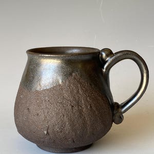 May include: A ceramic mug with a rounded body featuring a dual-texture finish. The upper portion and handle have a smooth, metallic dark bronze glaze, while the lower section displays a rough, dark brown, unglazed, and textured clay surface.