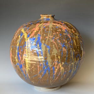 May include: A large, spherical ceramic vase with a brown base and colourful paint splatters in blue, yellow, pink, and gold. The vase has a narrow neck and sits on a small, white base. The design is abstract and artistic.
