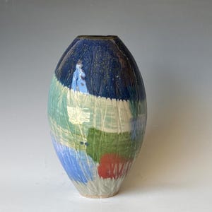 May include: An oval-shaped ceramic vase featuring a textured surface with vertical carved lines. The glaze displays a colour palette of deep blue at the top, transitioning into bands of light green, white, bright blue, and a patch of red near the base.