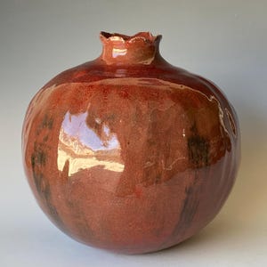 May include: A handcrafted ceramic vase in the shape of a pomegranate. The vase features a glossy, reddish-brown glaze with darker, mottled accents. The top has a ruffled, textured rim.