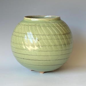 May include: A round, pale green ceramic vase with a textured surface and horizontal gray lines. The vase has a small opening at the top and rests on three small feet. The overall design is simple and elegant.