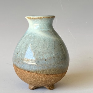 May include: A small, round ceramic vase featuring a light blue, glossy glaze on the upper portion and a textured, unglazed brown clay base. The vessel has a narrow neck with a flared rim and stands on three small, integrated clay feet.