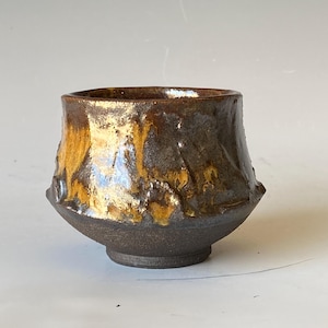 May include: A handcrafted ceramic bowl with a unique, textured surface. The bowl features a dark brown base that transitions to a golden-brown and bronze upper section. The bowl's design is organic and rustic.