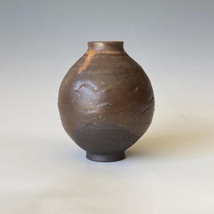 May include: A small, handcrafted brown ceramic vase with a rounded body and a narrow neck. The vase has a textured surface with darker tones at the base and a lighter brown at the top. The vase is approximately 10 cm tall.
