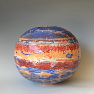 May include: A spherical ceramic vase with a textured surface. The vase is painted with horizontal bands of blue, red, and orange, with white and blue paint splatters. The top is a darker blue.