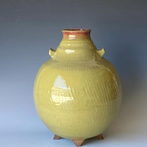 May include: A round ceramic vase with a glossy yellow glaze, featuring a narrow neck with a reddish-orange rim, two small side handles, and three short, textured brown feet at the base. The surface has a subtle, repeating diagonal pattern.