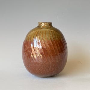 May include: A handcrafted ceramic vase with a rounded body and a narrow neck. The vase features a spiral design and a gradient of colors, transitioning from a reddish-brown base to a golden-brown top. The vase has a glossy finish.
