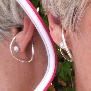 May include: A woman with short, silver hair is wearing a pair of silver hoop earrings with a pearl accent. The earrings are visible in the reflection of a mirror.