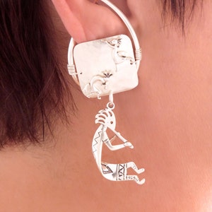 May include: Silver ear cuff with a dangling charm of a Native American flute player. The charm is attached to a square, hammered silver plate with a design of birds and flowers. The ear cuff is worn on the left ear.