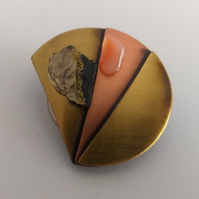 Peut inclure: Une broche en laiton, cuivre et m&eacute;tal noir de forme semi-circulaire. La broche pr&eacute;sente un petit morceau de pierre brun clair avec des paillettes dor&eacute;es et une pierre rose.