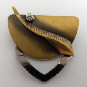 Peut inclure: Une broche en m&eacute;tal dor&eacute; avec un motif de feuille stylis&eacute; et un strass transparent. La broche a une finition textur&eacute;e et une forme g&eacute;om&eacute;trique.