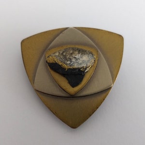 Peut inclure: Un pendentif triangulaire avec un design en couches. La couche centrale est en m&eacute;tal dor&eacute; avec une pierre noire et blanche au centre. Les couches externes sont en m&eacute;tal argent&eacute;.