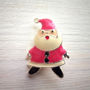 Pode incluir: Um broche de Papai Noel vermelho e branco com barba branca e olhos pretos. O broche tem um contorno prateado e um chapéu vermelho com um pompom branco.