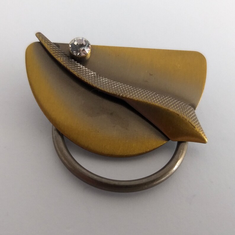Peut inclure: Une broche de couleur or et argent avec un motif g&eacute;om&eacute;trique et un strass transparent. La broche a une forme incurv&eacute;e et une bague attach&eacute;e au bas.