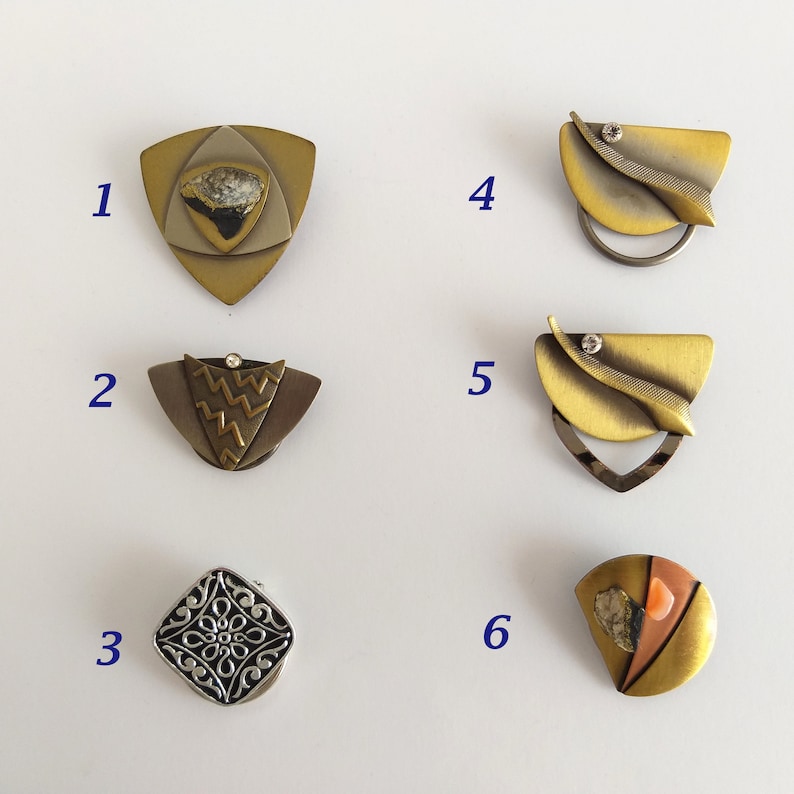 Peut inclure: Six broches en m&eacute;tal diff&eacute;rentes avec des motifs et des couleurs vari&eacute;s. Les broches sont dispos&eacute;es en grille sur un fond blanc.