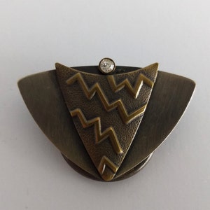 Peut inclure: Une broche de couleur bronze et or avec un motif g&eacute;om&eacute;trique et un strass transparent au centre.