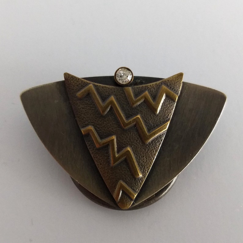 Peut inclure: Une broche de couleur bronze et or avec un motif g&eacute;om&eacute;trique et un strass transparent au centre.