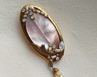 May include: An oval-shaped vintage brooch with a mother-of-pearl center. The gold-toned frame is adorned with small pearls and iridescent crystals. A decorative dangle with a crystal pendant hangs from the bottom.