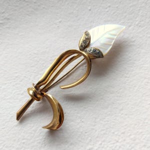 Puede incluir: Un broche de metal dorado con forma de tallo estilizado y un lazo curvado. La parte superior presenta una incrustación blanca e iridiscente en forma de hoja, acentuada por dos pequeñas secciones plateadas con piedras redondas brillantes.