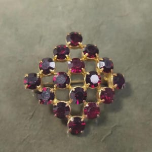 Può includere: Spilla vintage dal design geometrico. La spilla presenta una montatura in metallo dorato incastonata con numerose pietre preziose rotonde di colore rosso scuro. Le pietre sono disposte a forma di diamante, creando un accessorio scintillante e accattivante.