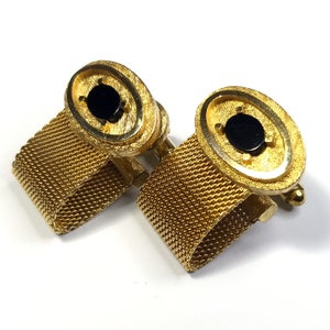 Könnte beinhalten: Ein Paar goldene Manschettenknöpfe mit schwarzen ovalen Einsätzen. Die Manschettenknöpfe sind an einem goldenen Maschenband befestigt.