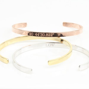 May include: Three stacked bracelets, one gold, one silver, and one rose gold. The rose gold bracelet has the coordinates "N 40 44'30.822" W" engraved on it.