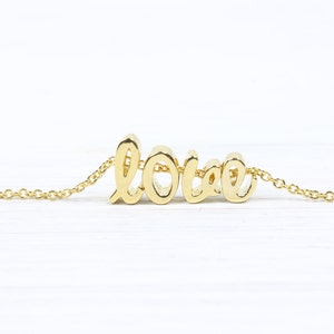 Könnte beinhalten: Eine goldene Halskette mit einem Anhänger in Form des Wortes 'love' in Kursivschrift.