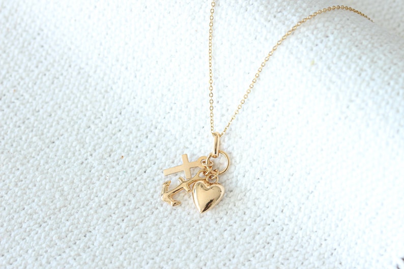 May include: A gold necklace with a heart, cross, and anchor charm. The necklace is on a white fabric background.