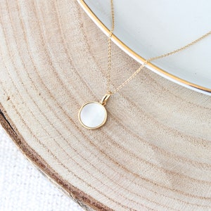 May include: A gold chain necklace with a white round pendant. The pendant is set in a gold frame and has a smooth, slightly textured surface.