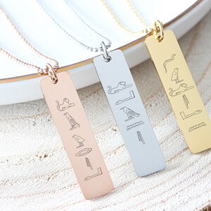 May include: Three gold, silver, and rose gold necklaces with a rectangular pendant featuring an engraved design of ancient Egyptian hieroglyphs.