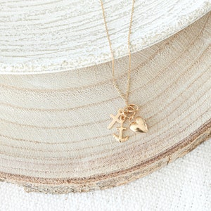 May include: Gold necklace with a heart, anchor, and cross charm. The necklace is on a wooden surface.