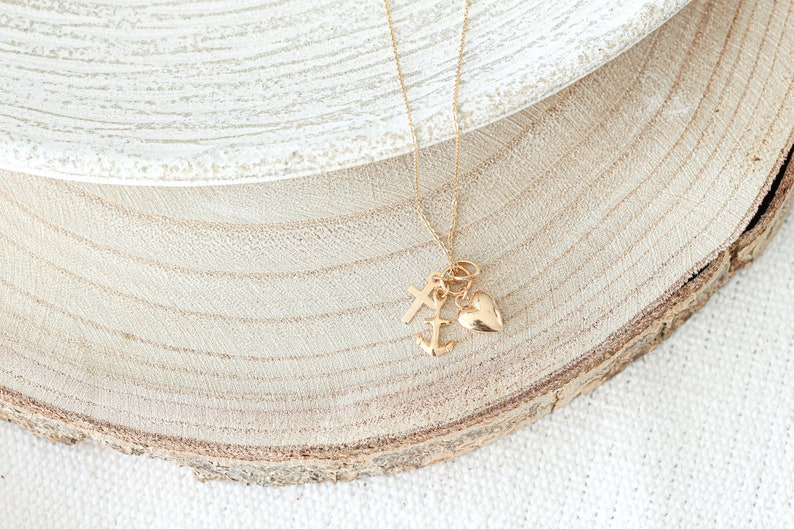 May include: Gold necklace with a heart, anchor, and cross charm. The necklace is on a wooden surface.