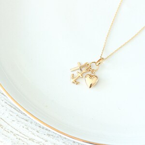 May include: A gold necklace with three charms: a cross, an anchor, and a heart. The charms hang from a fine gold chain. The necklace is presented on a white surface with a gold trim.