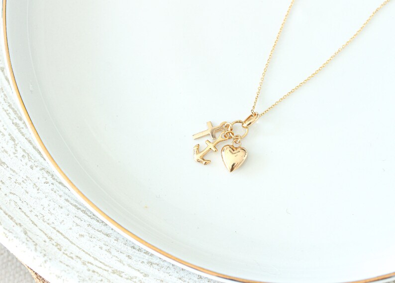 May include: A gold necklace with three charms: a cross, an anchor, and a heart. The charms hang from a fine gold chain. The necklace is presented on a white surface with a gold trim.