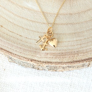 May include: Gold necklace with a heart, cross, and anchor charm. The necklace is on a wooden surface.