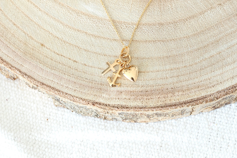 May include: Gold necklace with a heart, cross, and anchor charm. The necklace is on a wooden surface.