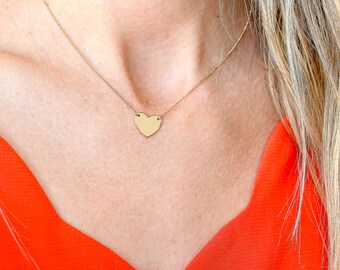 May include: A gold heart pendant necklace on a woman's neck. The necklace is on a chain and the heart is a simple, flat design.