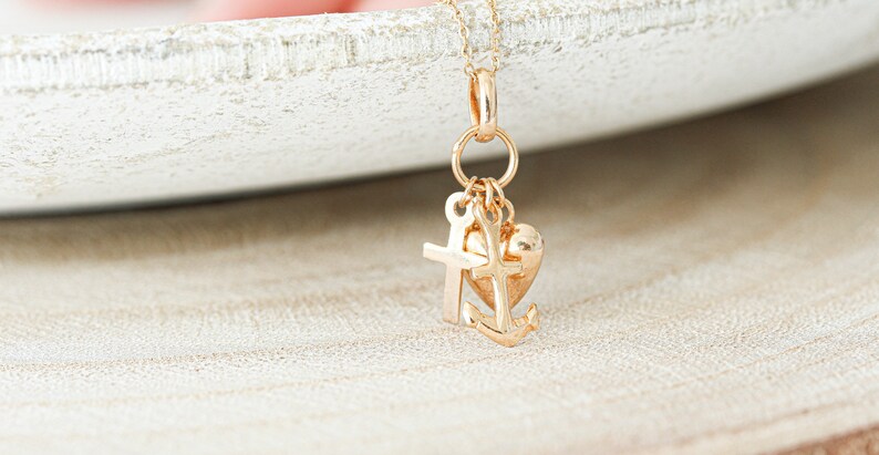 May include: Gold charm necklace with a heart, cross, and anchor pendant. The charms are attached to a gold chain.