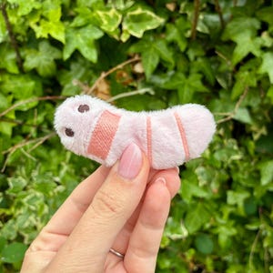 May include: A small, plush, light pink caterpillar-shaped toy with two dark brown embroidered eyes and two horizontal coral-coloured stitched bands across its body. The soft, fuzzy texture is visible on the surface of the segmented, curved form.