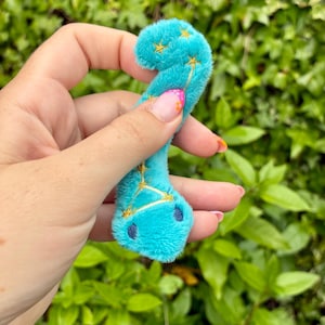 May include: A small, bright turquoise plush toy shaped like a curved worm or snake. The soft, fuzzy material features gold embroidered star patterns and constellations along its body, with two dark blue embroidered eyes at the rounded end.